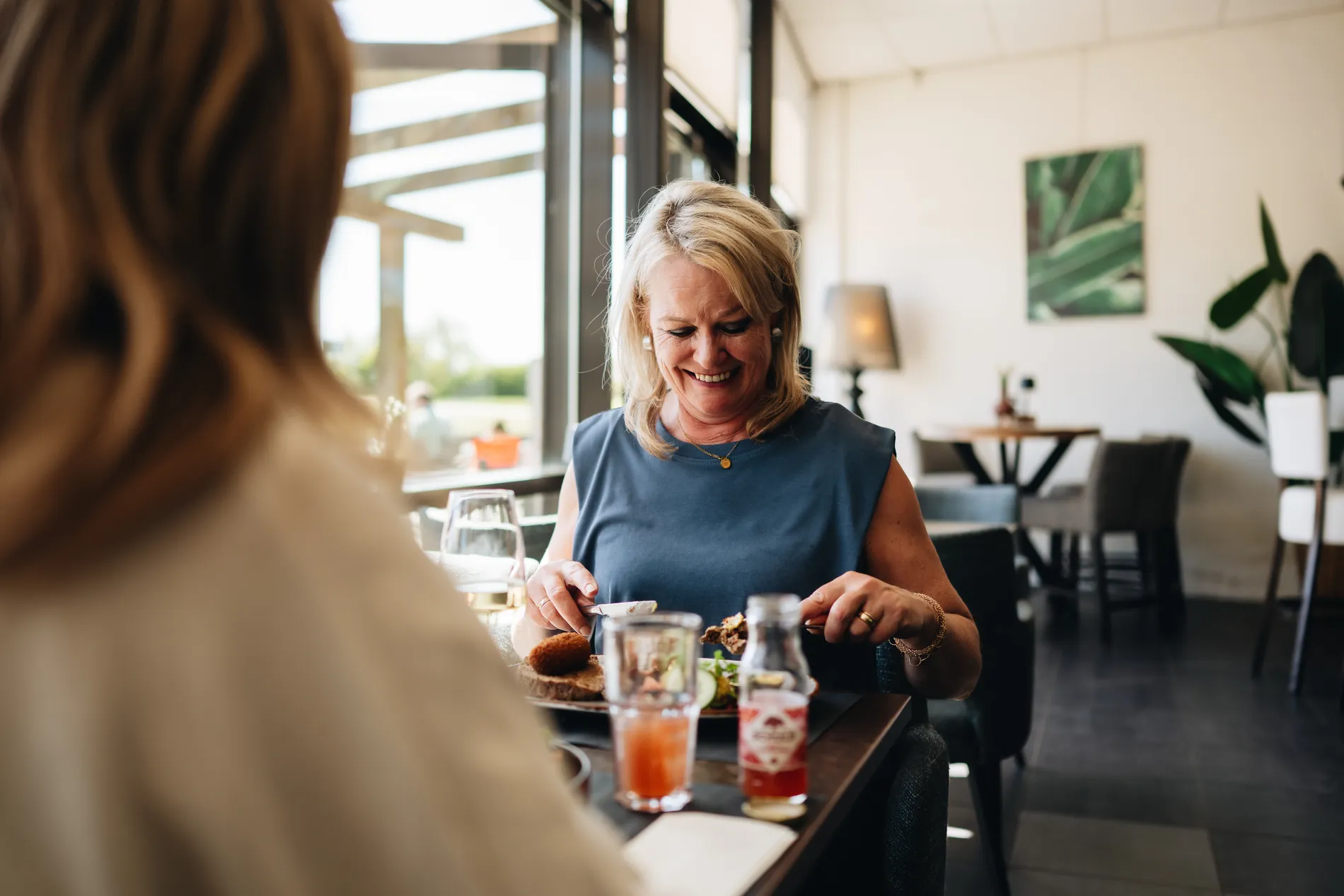 Een glimlachende blonde vrouw die geniet van haar lunch binnen in het restaurant, terwijl ze haar gerecht met mes en vork aansnijdt.